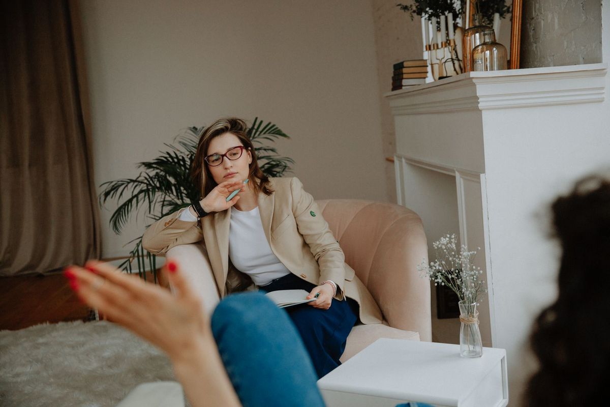 A counselor in glasses and a tan blazer sits in a pink armchair, listening attentively as a person in the foreground talks and gestures with her hand.